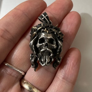 May include: Silver pendant featuring a skull with a crown of thorns and a beard. The skull is detailed with a cross on the forehead and a gold accent on the crown.