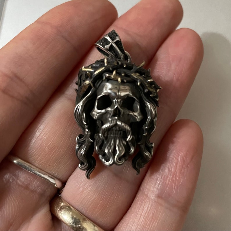 May include: Silver pendant featuring a skull with a crown of thorns and a beard. The skull is detailed with a cross on the forehead and a gold accent on the crown.