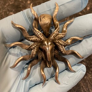 May include: A silver-toned pendant featuring a detailed skull with horns and a sunburst design. The pendant has a textured surface and intricate details.