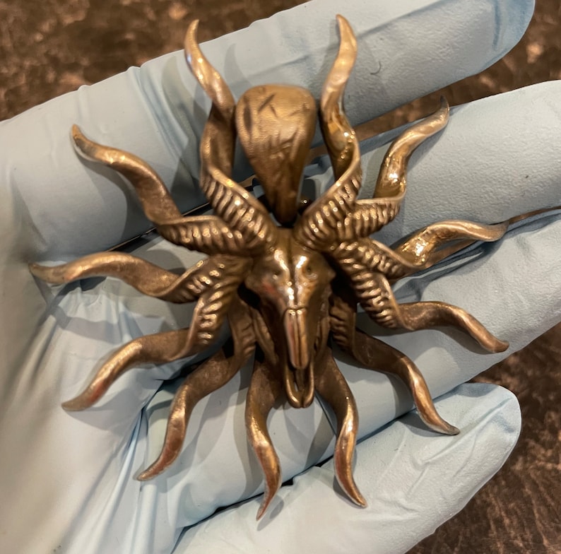 May include: A silver-toned pendant featuring a detailed skull with horns and a sunburst design. The pendant has a textured surface and intricate details.