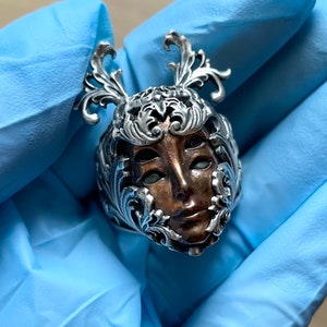 May include: A silver ring with a detailed design of a face with ornate floral accents. The face is brown and has a detailed design.