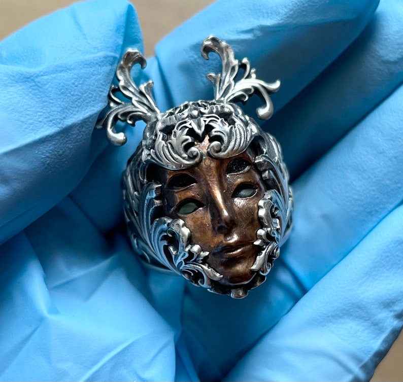May include: A silver ring with a detailed design of a face with ornate floral accents. The face is brown and has a detailed design.