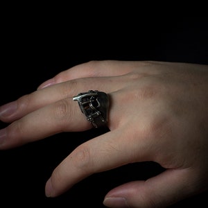 May include: A silver ring with a detailed design of a buckle and straps. The ring is worn on a person's finger.