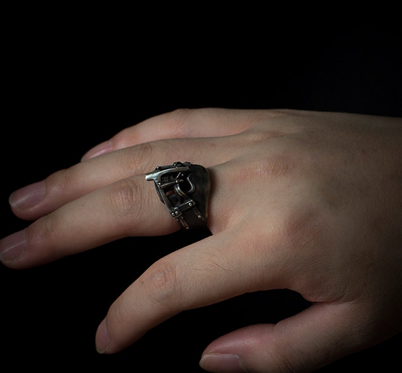 May include: A silver ring with a detailed design of a buckle and straps. The ring is worn on a person's finger.