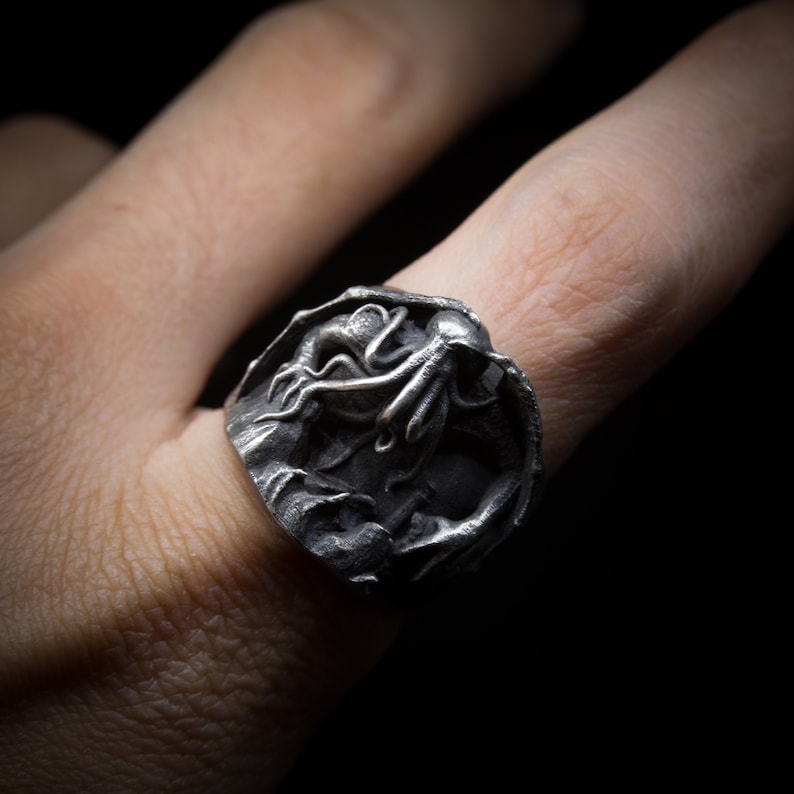 May include: A silver ring with a detailed sculpture of a tentacled creature, possibly an octopus or squid, emerging from a rocky surface.