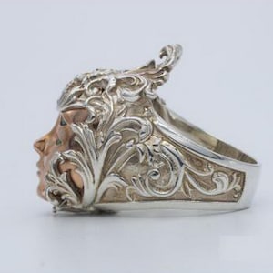May include: A silver ring with a detailed design of a face with a floral crown. The face is made of rose gold.