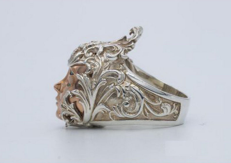 May include: A silver ring with a detailed design of a face with a floral crown. The face is made of rose gold.