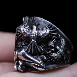 May include: A silver ring with a detailed design of a bird with outstretched wings, a skull, and a human figure. The ring is worn on a finger.