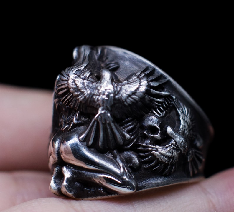 May include: A silver ring with a detailed design of a bird with outstretched wings, a skull, and a human figure. The ring is worn on a finger.