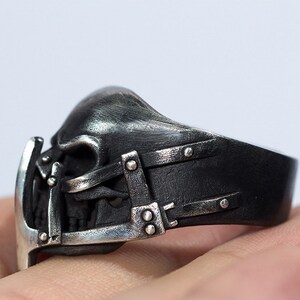 May include: A silver ring with a skull design. The skull is detailed with a metal cage and straps. The ring is worn on a finger.