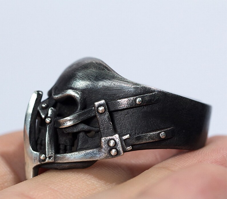 May include: A silver ring with a skull design. The skull is detailed with a metal cage and straps. The ring is worn on a finger.