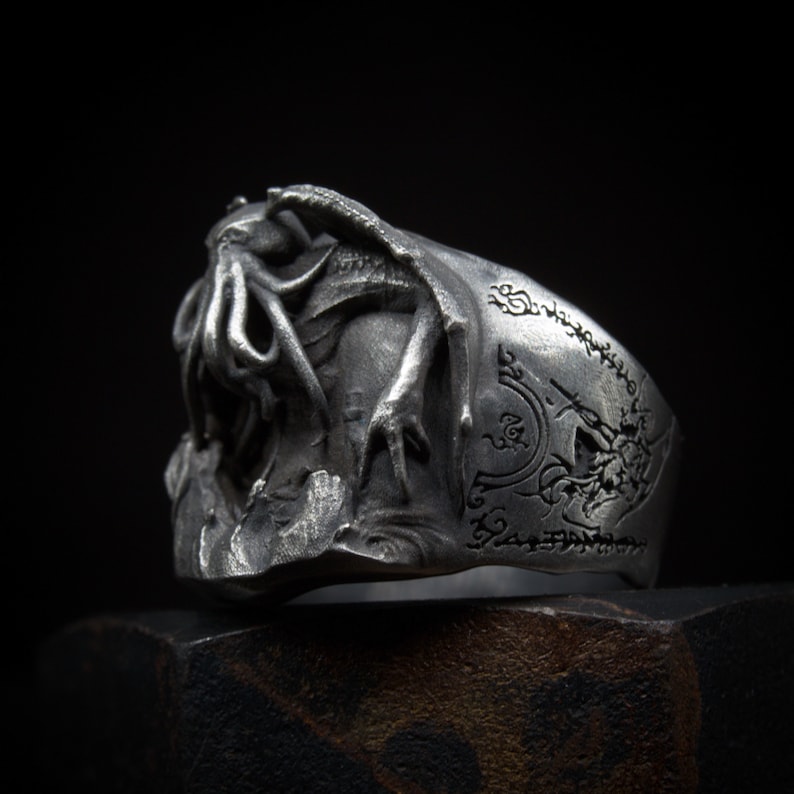 May include: A silver ring with a detailed design of a creature with tentacles and claws. The ring band is engraved with a circular design and script.