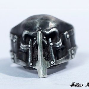 May include: A silver skull ring with a detailed design. The skull has a pointed nose and a metal bar across the mouth. The ring is a statement piece for any jewelry collection.