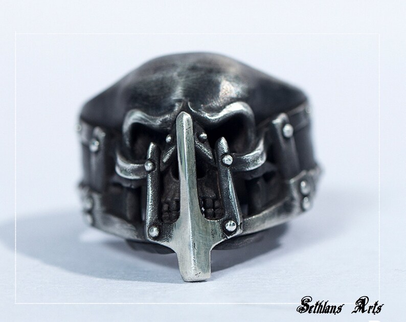 May include: A silver skull ring with a detailed design. The skull has a pointed nose and a metal bar across the mouth. The ring is a statement piece for any jewelry collection.