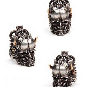 Puede incluir: Un anillo de plata y oro con un dise&ntilde;o detallado de la cara de un demonio. El anillo tiene ojos verdes y cuernos.