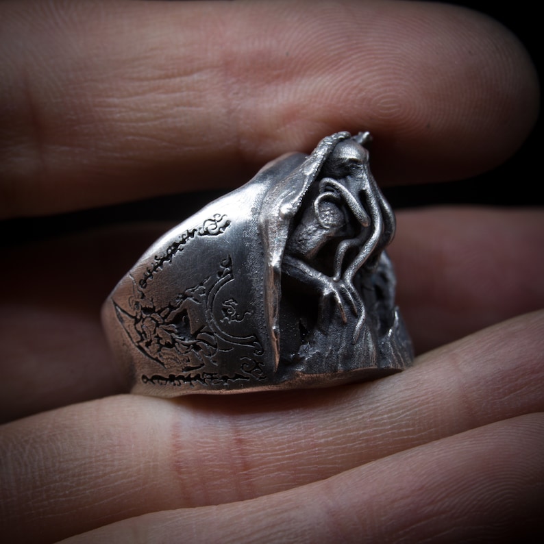 May include: A silver ring with a detailed design of an octopus-like creature with tentacles. The ring is worn on a finger and has intricate details.