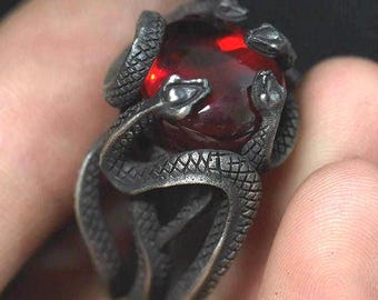 May include: A silver ring with a red gemstone surrounded by three intertwined snake designs.