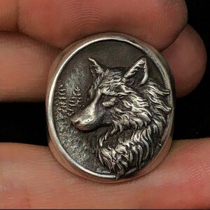 Op de afbeelding: Een zilveren ring met een ovale voorkant met een gedetailleerd reliëf van een wolfskop in profiel, tegen een achtergrond van gestileerde naaldbomen. De ring heeft een geborstelde metalen afwerking, wat hem een vintage uitstraling geeft.