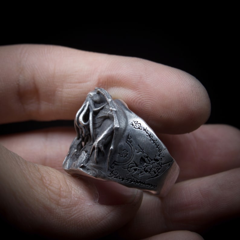May include: A silver ring with a detailed, three-dimensional design of a creature with tentacles. The ring band is inscribed with a series of symbols.
