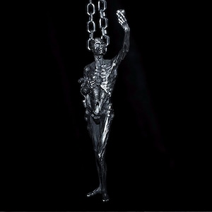Op de afbeelding: Een zilverkleurige sculptuur van een skeletfiguur, opgehangen aan kettingen aan het hoofd. De figuur houdt een voorwerp omhoog in zijn rechterhand. De sculptuur heeft een gedetailleerde ribbenkast en staat tegen een zwarte achtergrond.