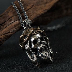 May include: Silver pendant necklace featuring a detailed skull with a crown of thorns. The skull has a long beard and flowing hair. The pendant is on a silver chain.