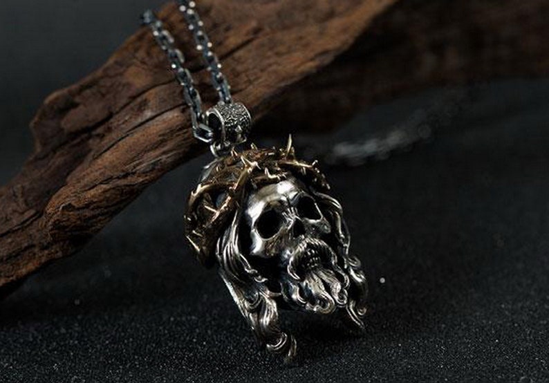 May include: Silver pendant necklace featuring a detailed skull with a crown of thorns. The skull has a long beard and flowing hair. The pendant is on a silver chain.