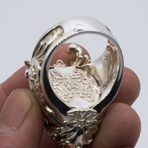 May include: A silver ring with intricate details and a large, ornate design. The ring is worn on a finger and the inside of the band is visible.