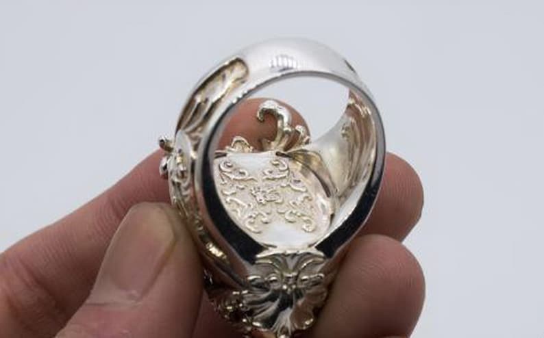May include: A silver ring with intricate details and a large, ornate design. The ring is worn on a finger and the inside of the band is visible.