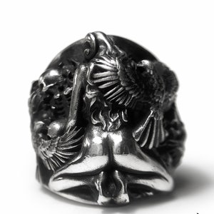 May include: A silver ring with a detailed design of a woman with wings. The woman is depicted in a seated position with her wings spread out behind her.