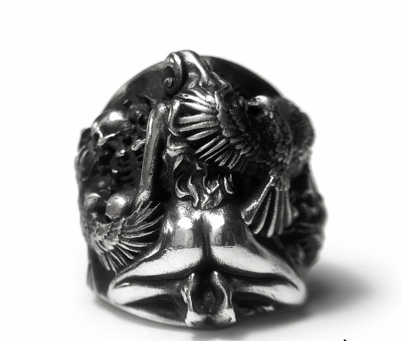 May include: A silver ring with a detailed design of a woman with wings. The woman is depicted in a seated position with her wings spread out behind her.