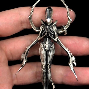 May include: A silver-toned pendant featuring a biomechanical creature with an elongated head, ribbed torso, and multiple limbs. The figure is positioned with hands together in a prayer-like gesture, framed by a circular, segmented arching structure.