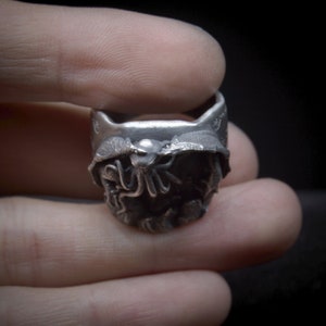 May include: A silver ring with a detailed design of a creature with tentacles. The ring is worn on a finger.