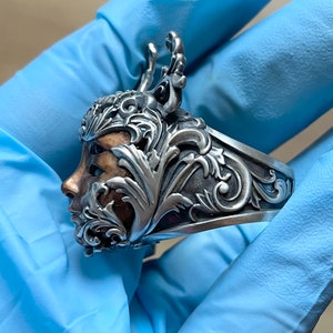 May include: A silver ring with a detailed design of a face surrounded by ornate leaves and branches. The face is partially obscured by the leaves, but the eye and nose are visible. The ring is made of silver and has a polished finish.