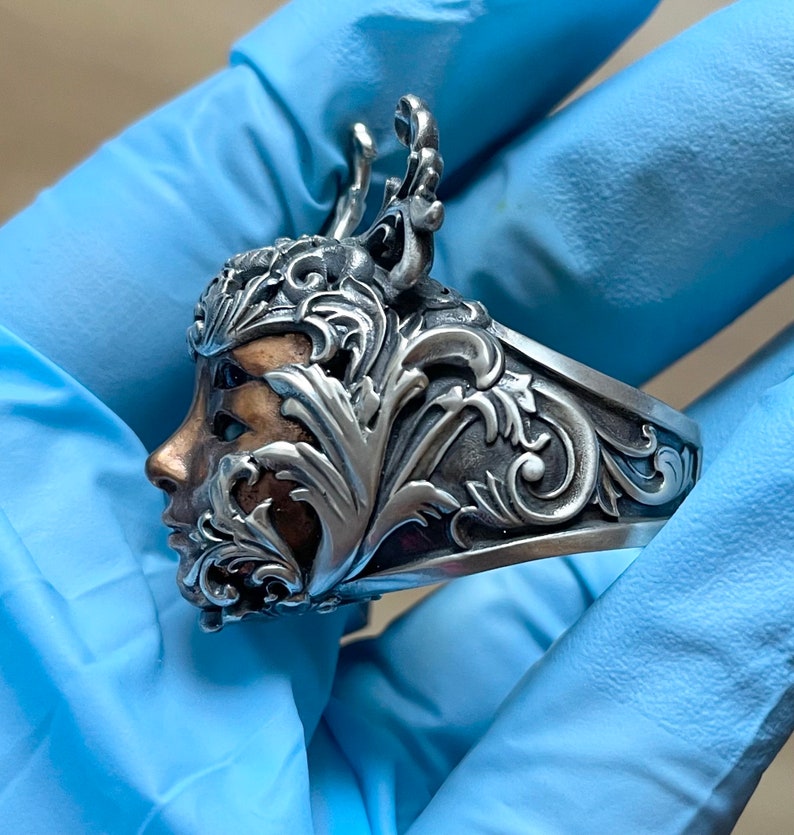 May include: A silver ring with a detailed design of a face surrounded by ornate leaves and branches. The face is partially obscured by the leaves, but the eye and nose are visible. The ring is made of silver and has a polished finish.