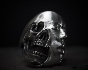May include: A silver ring with a detailed skull and human face design. The skull is open and the face is closed with the eyes shut. The ring is made of metal and has a smooth finish.