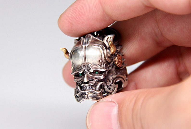 Puede incluir: Un anillo de plata y oro con un dise&ntilde;o detallado de la cara de un demonio. El demonio tiene ojos verdes y cuernos. El anillo se lleva en un dedo.