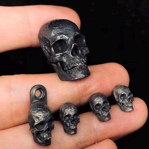 Könnte beinhalten: Ein Set aus fünf schwarzen Metall-Schädelanhängern in verschiedenen Größen. Der größte Schädel ist etwa 5 cm hoch und hat ein detailliertes Design. Die kleineren Schädel sind etwa 2,5 cm hoch und haben ein einfacheres Design.
