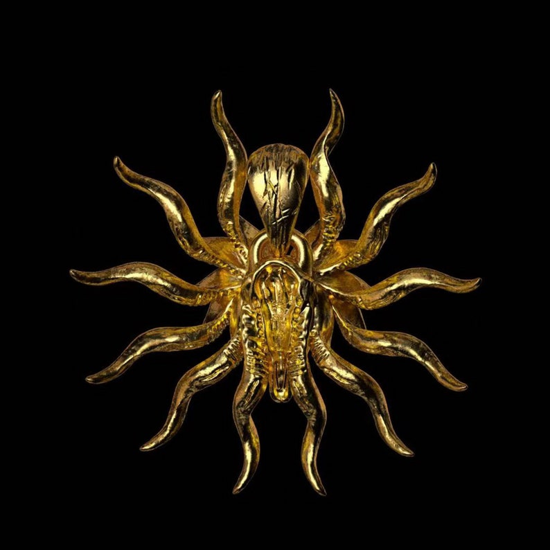 May include: A gold-colored pendant in the shape of a stylized sun with multiple rays. The pendant has a detailed design and a textured surface.