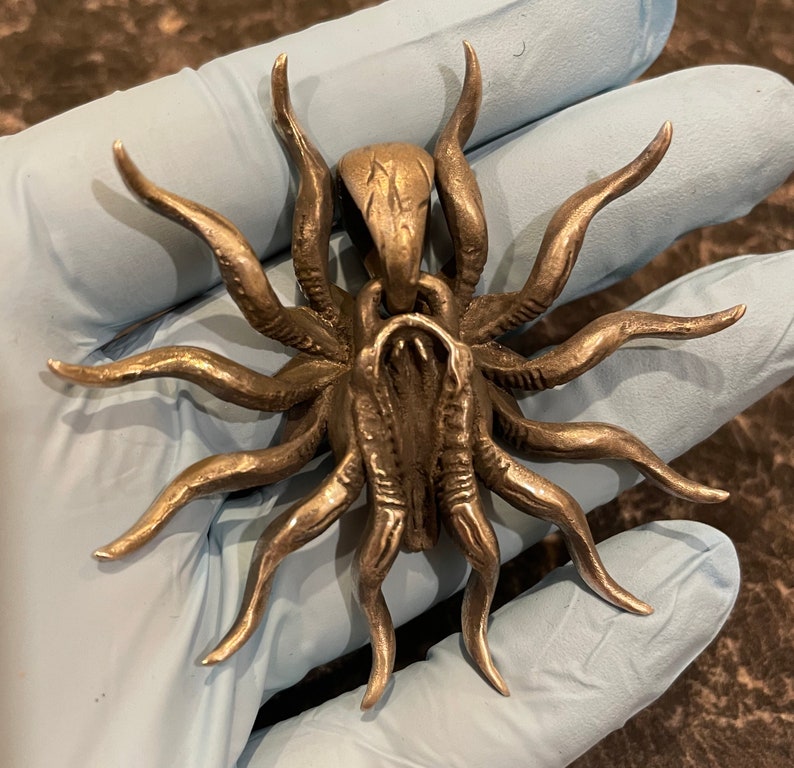 May include: A silver pendant in the shape of a spider with eight legs. The spider is detailed with textured lines and a raised, textured body.