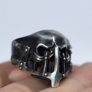 May include: A silver skull ring with a detailed design. The skull has a cage-like structure around the face and a pointed nose.