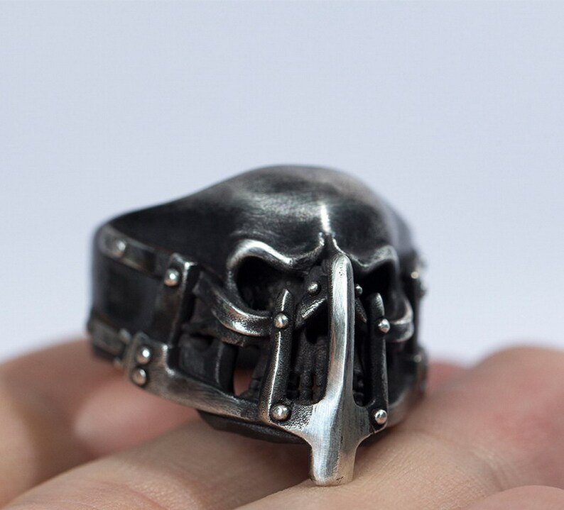 May include: A silver skull ring with a detailed design. The skull has a cage-like structure around the face and a pointed nose.