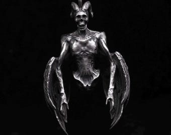 May include: A silver pendant depicting a skeletal figure with horns and wings. The figure has a detailed, textured surface and appears to be in a flying pose.