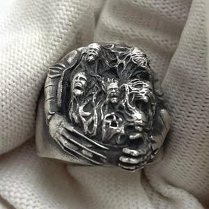 May include: A silver ring with a detailed design of multiple faces intertwined. The faces are sculpted with intricate details and appear to be in a state of distress or anguish.
