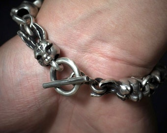 May include: A silver bracelet with a skull and crossbones design. The bracelet is made of thick, chunky links and has a toggle clasp.