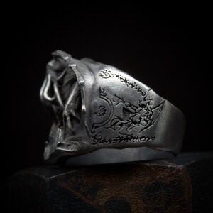 May include: A silver ring with a detailed design of a creature with wings and a skull. The ring is engraved with a pattern of runes.