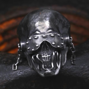Puede incluir: Un anillo de metal oscuro con forma de calavera que lleva un antifaz remachado sobre las cuencas de los ojos. La calavera presenta barras verticales que cubren la boca y pequeñas cadenas metálicas unidas a los lados, todo con acabado desgastado.