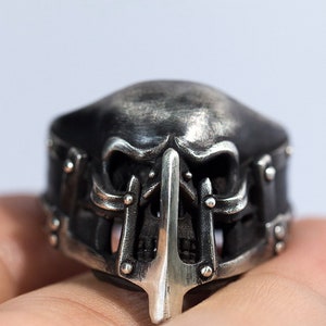 May include: A silver skull ring with a detailed design. The skull has a menacing expression with sharp teeth and a pointed nose. The ring is decorated with intricate metalwork and studs.