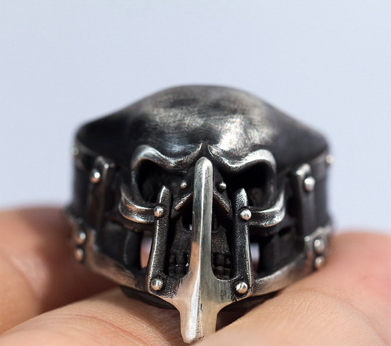 May include: A silver skull ring with a detailed design. The skull has a menacing expression with sharp teeth and a pointed nose. The ring is decorated with intricate metalwork and studs.