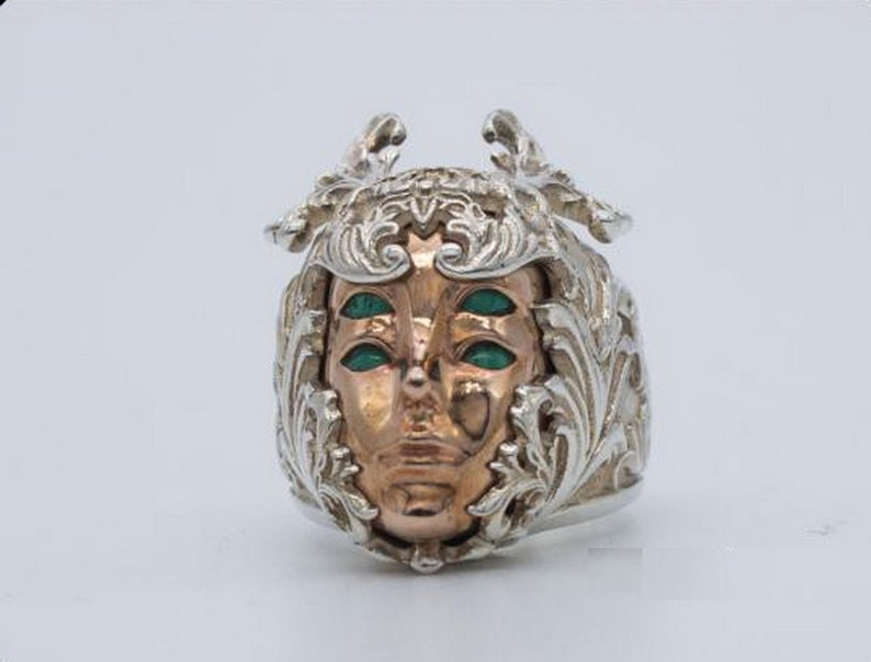May include: A silver ring with a detailed, ornate design featuring a face with three green eyes. The face is made of copper and is set within the silver design.