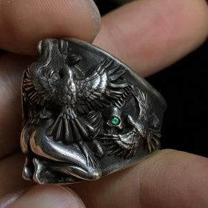 May include: A silver ring with a detailed design of a woman and two birds. The ring features a green gemstone.